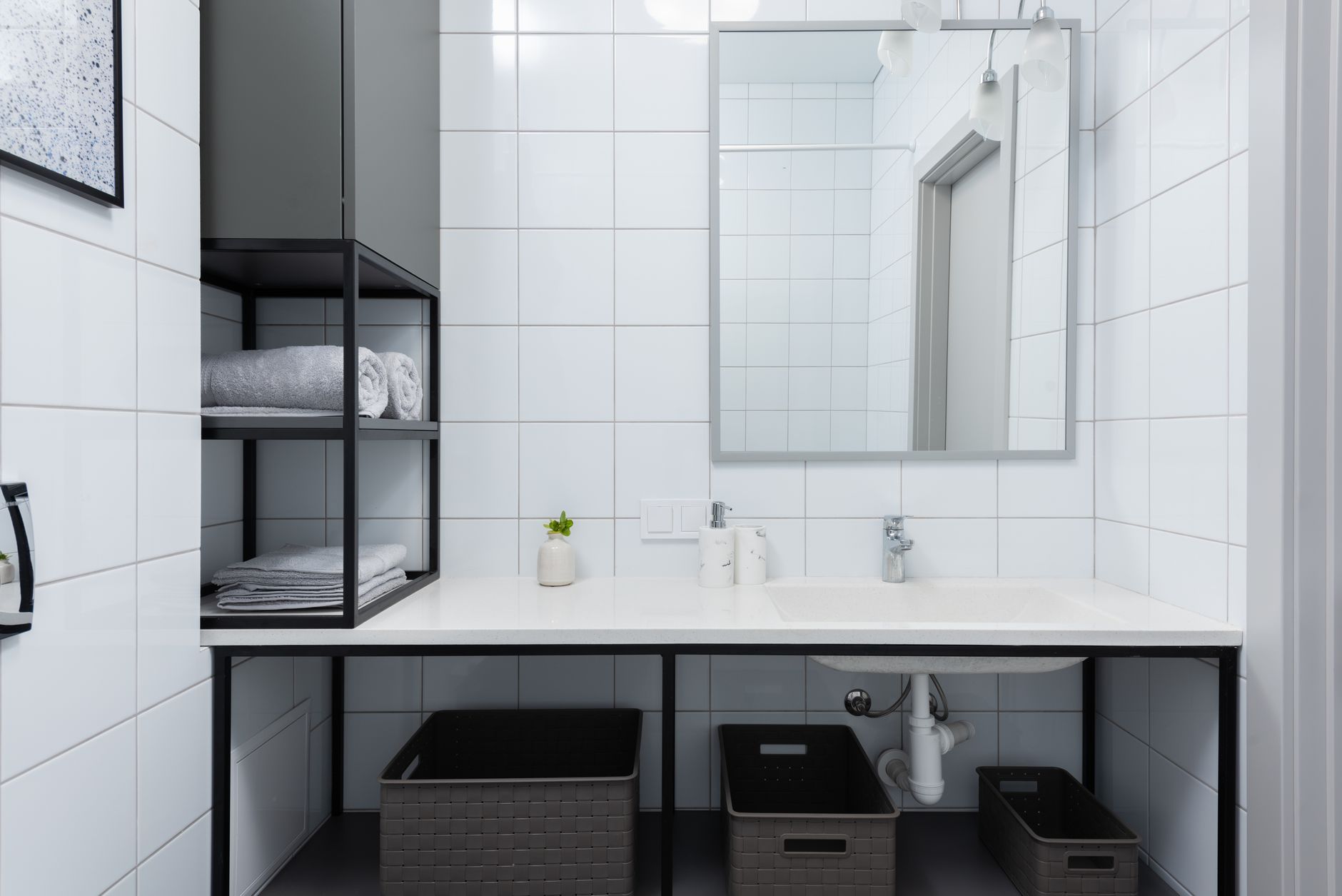 Interior of light bathroom with white counter near shelves with containers and towels near sink with faucet and mirror on tile near soap dispenser