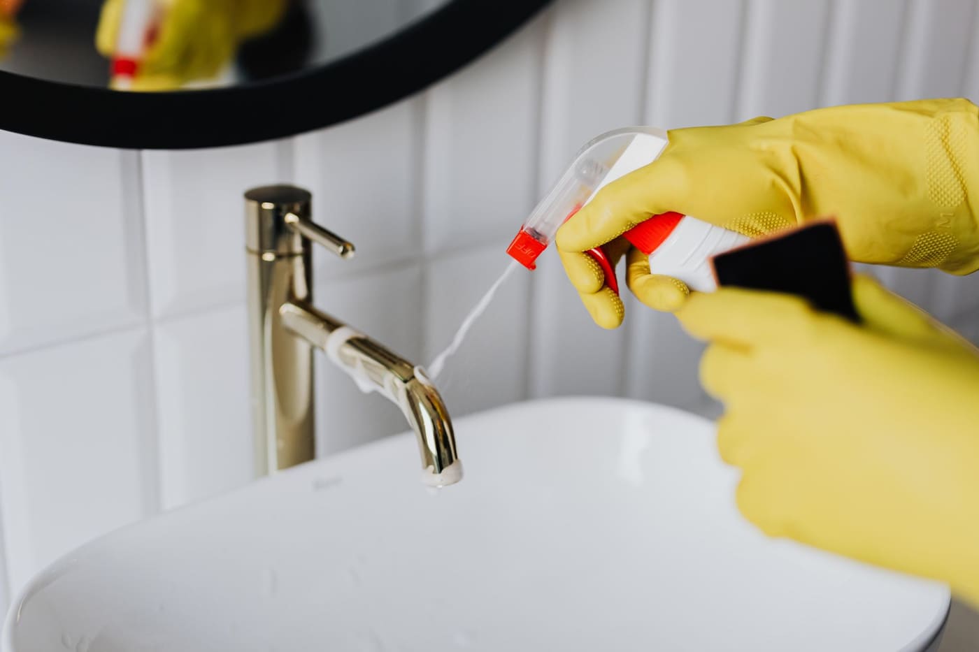 Gloved hands holding a spray bottle and cloth beside a bathroom faucet