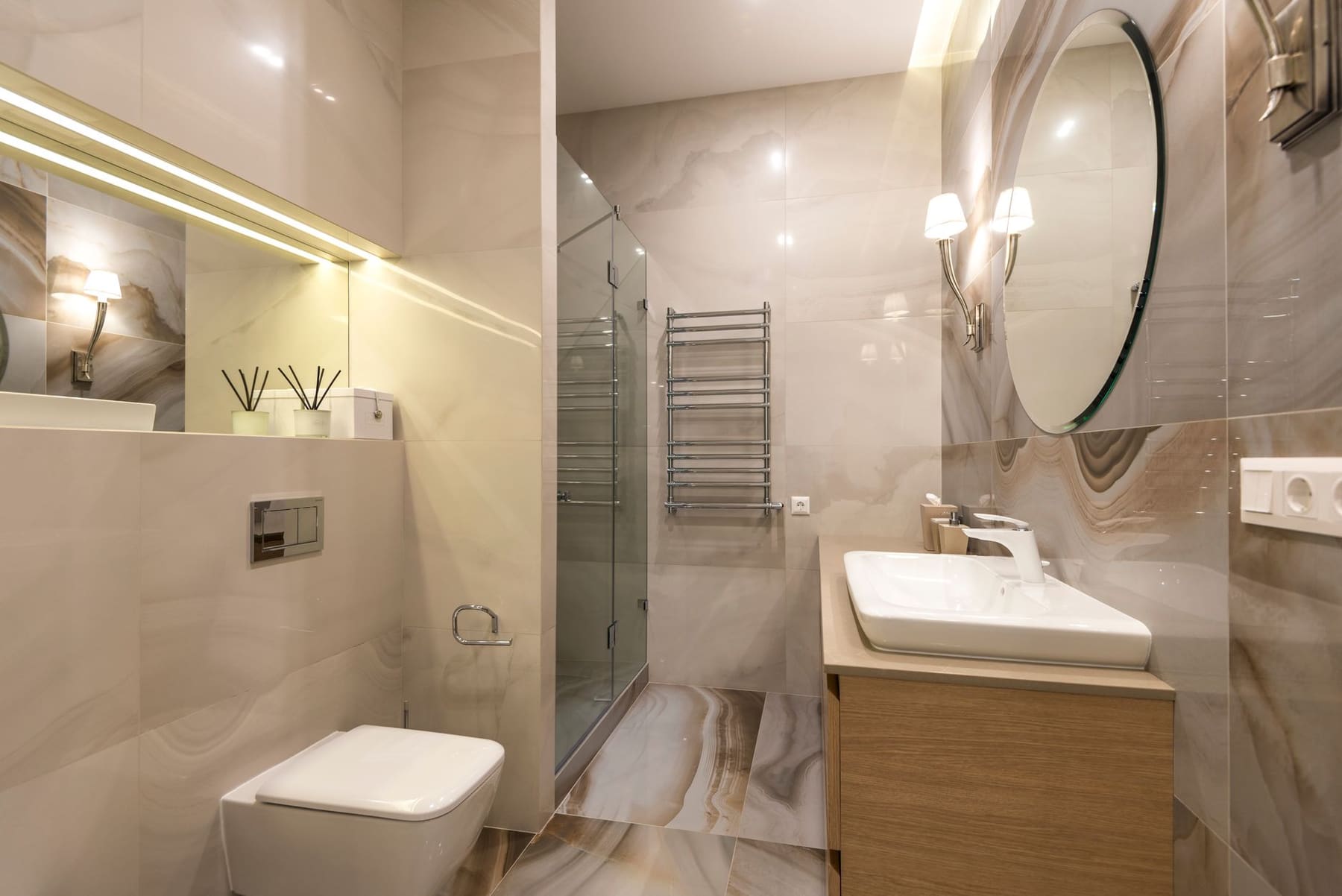 A bright bathroom with a round mirror, wall lights and a warm wood vanity