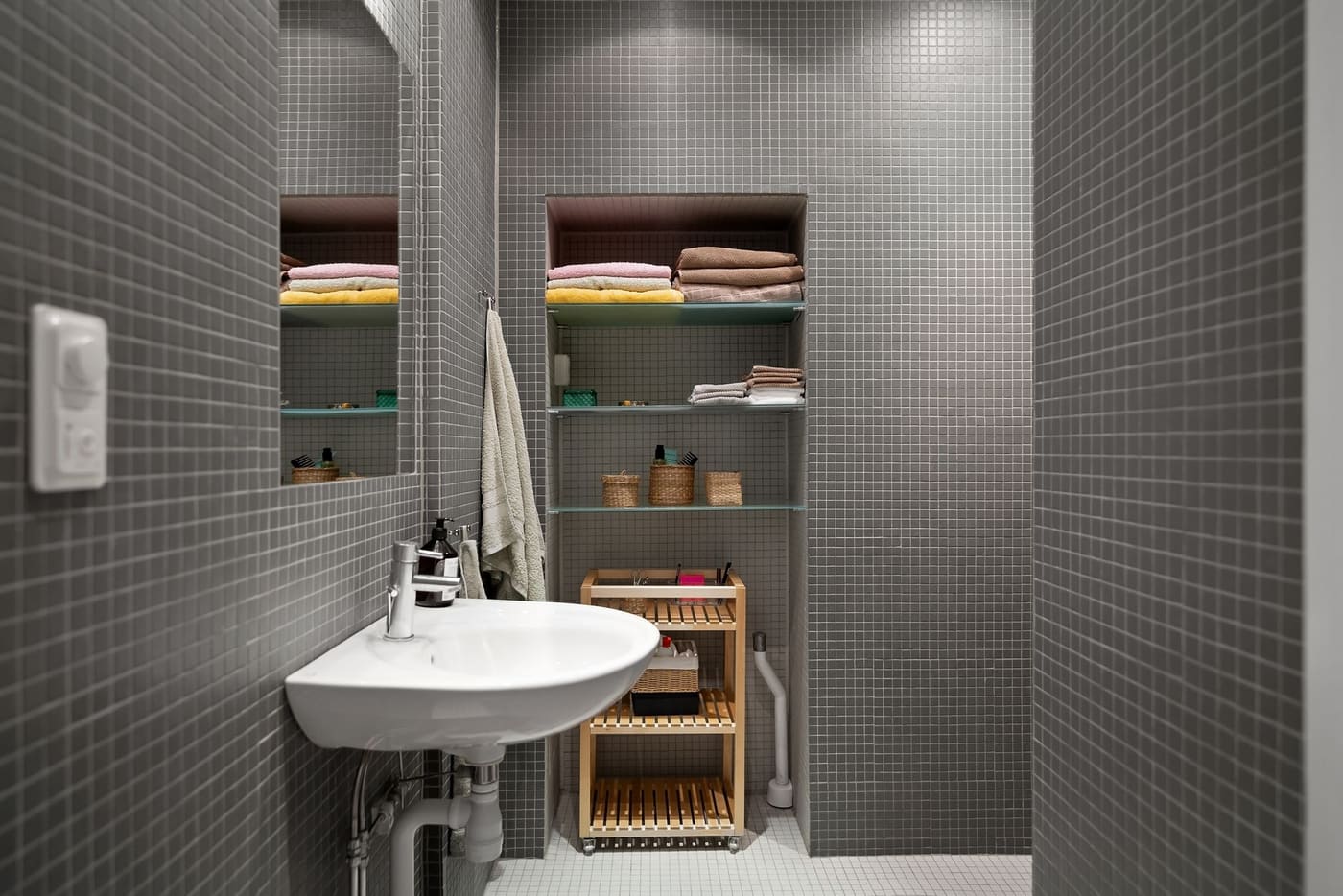 Small gray-tiled bathroom with towels on recessed shelves above a wall-mounted sink
