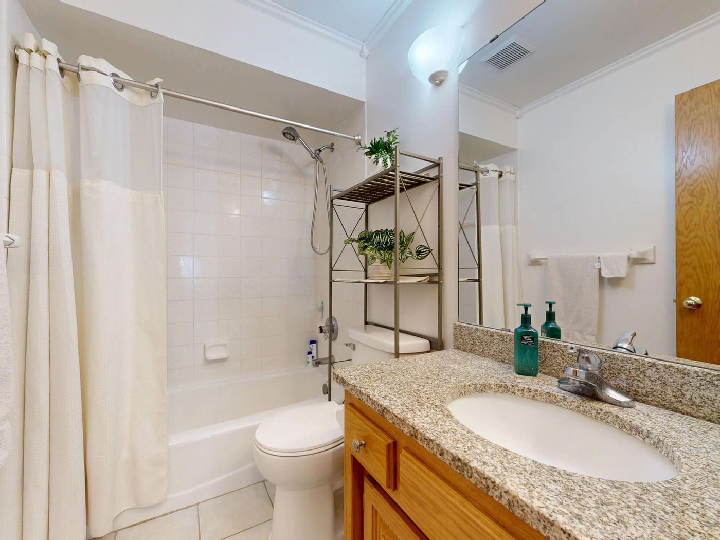 Bathroom with a freestanding metal shelf over the toilet, a cream shower curtain and a granite vanity